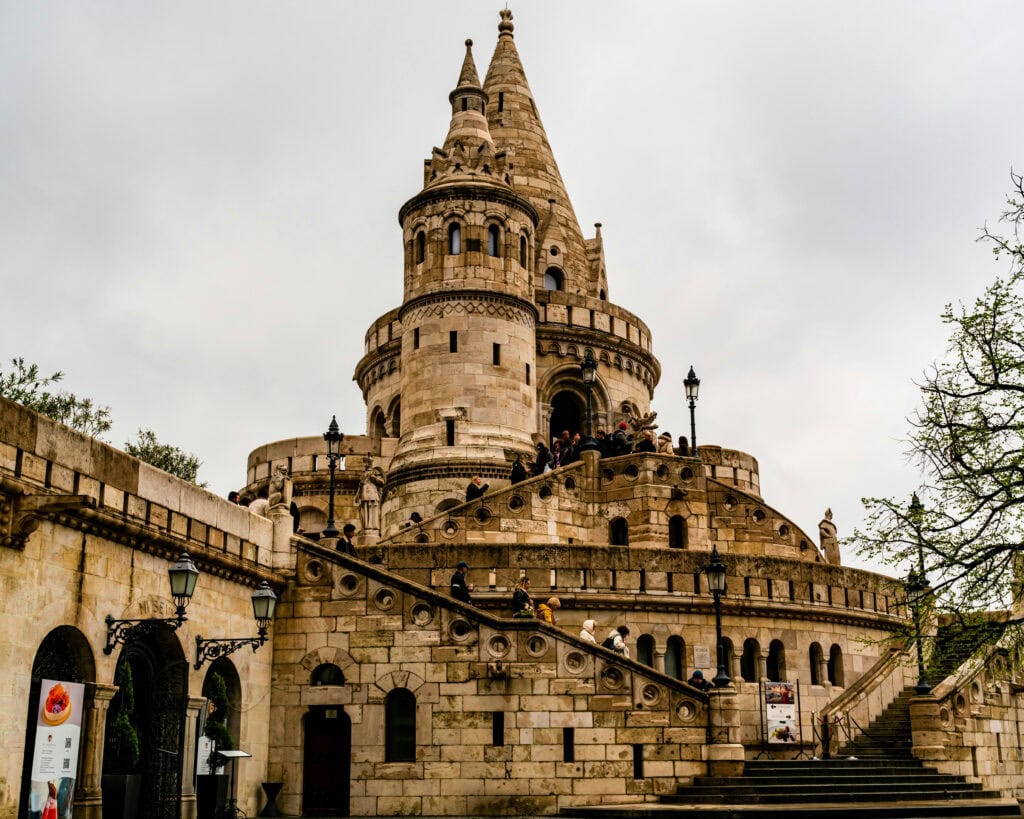Fisherman’s Bastion on Castle Hill in Budapest, showing pale stone terraces, staircases, and rounded towers with visitors climbing the steps under a grey, overcast sky.