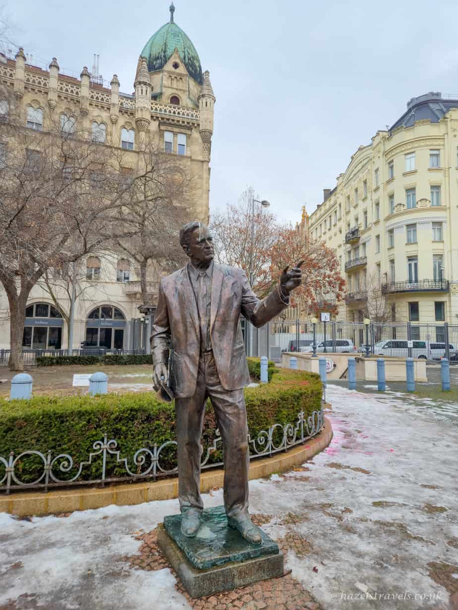Bronze statue of George H. W. Bush in Liberty Square, Budapest, with raised arm gesture, winter greenery and historic buildings in the background on a cold, overcast day.