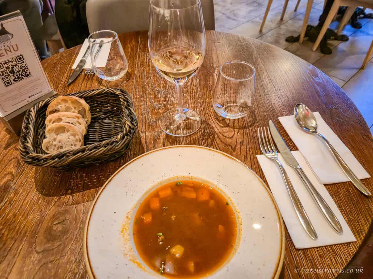 Bowl of Hungarian goulash soup served at a restaurant table, with rich brown broth and chunks of meat, bread and wine glasses visible alongside cutlery.