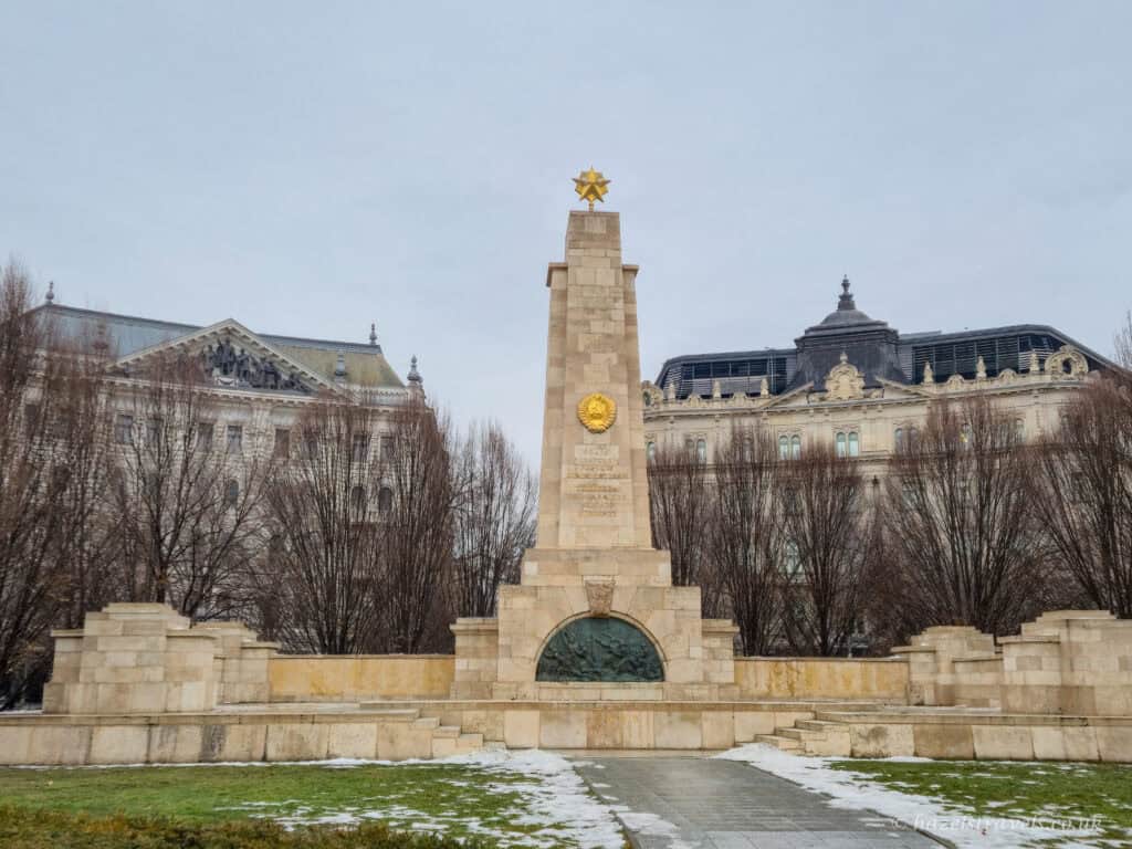 Soviet War Memorial in Liberty Square, Budapest, with a tall pale stone obelisk topped by a gold star, flanked by historic buildings, bare winter trees, and patches of snow on the grass under a pale grey sky.