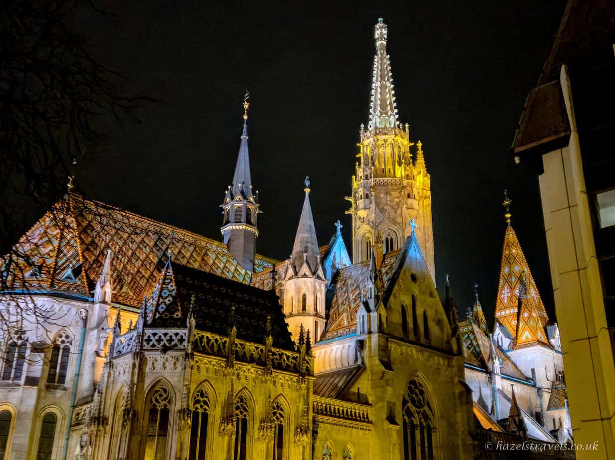 Matthias Church in Budapest at night, with colourful patterned roof tiles and tall Gothic spire lit in warm golden light against a dark sky.