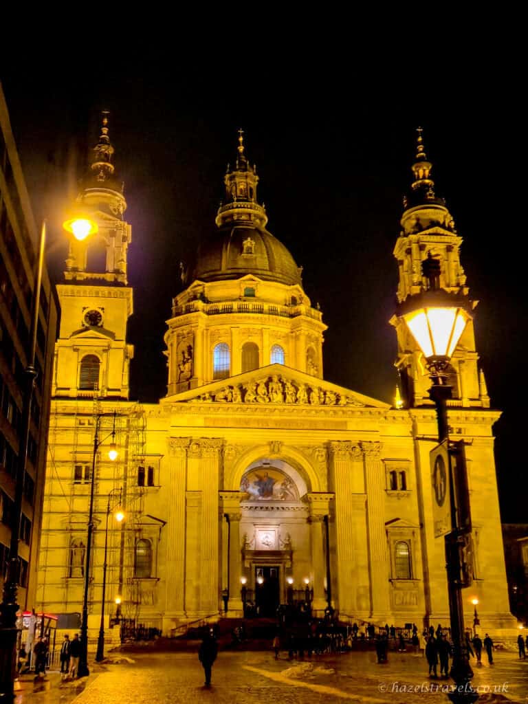 St. Stephen’s Basilica in Budapest at night, with twin bell towers and a large central dome glowing golden under floodlights, people gathered in the square below against a dark winter sky.