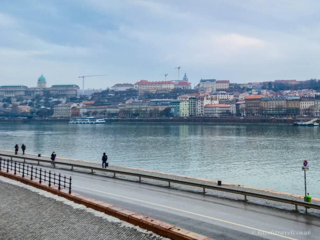 View across the Danube in Budapest during winter daylight, with riverside walkway and pedestrians in the foreground and pastel-coloured buildings and hills of Buda lining the opposite bank under a pale sky.