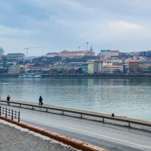 View across the Danube in Budapest during winter daylight, with riverside walkway and pedestrians in the foreground and pastel-coloured buildings and hills of Buda lining the opposite bank under a pale sky.