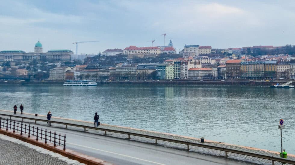 View across the Danube in Budapest during winter daylight, with riverside walkway and pedestrians in the foreground and pastel-coloured buildings and hills of Buda lining the opposite bank under a pale sky.