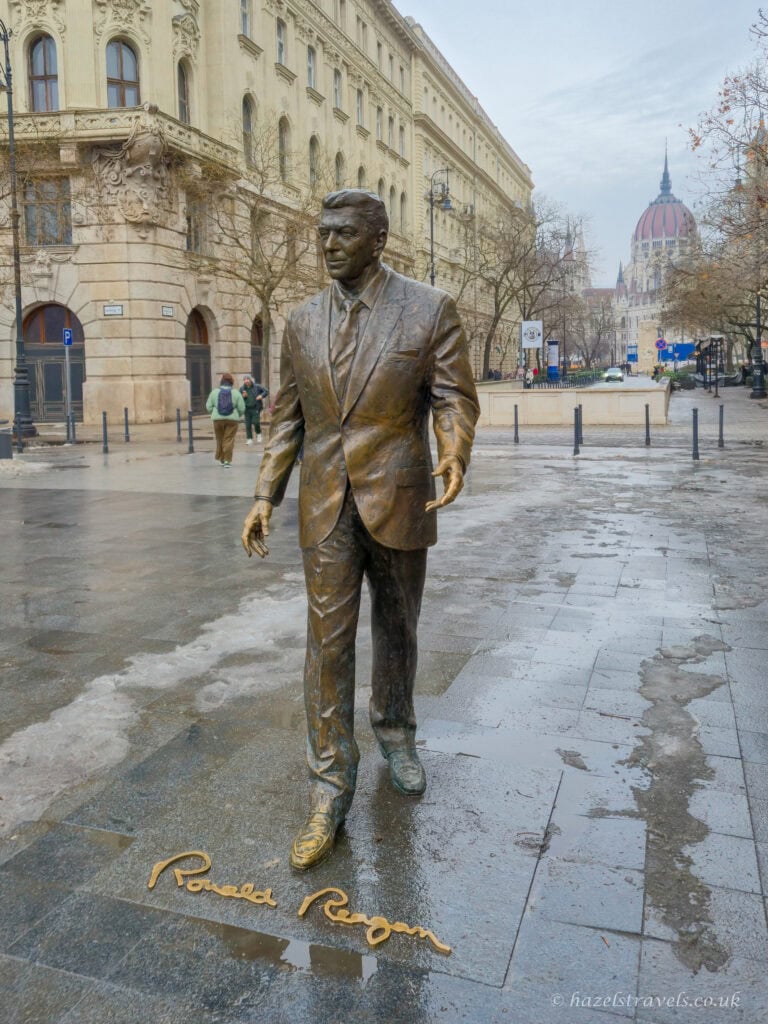 Bronze statue of Ronald Reagan in Budapest, standing on a wet pavement in Liberty Square, with historic stone buildings and the Hungarian Parliament dome visible in the background on a grey winter day.