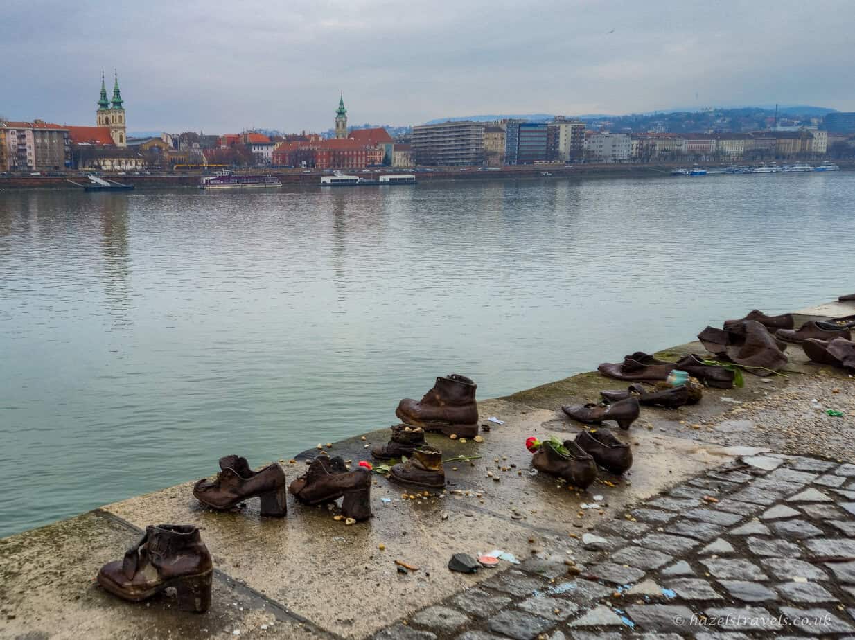 Shoes on the Danube Bank memorial in Budapest, with multiple rusted iron shoes lined along the river’s edge, small flowers placed inside some pairs, and the Danube flowing past under a grey sky.