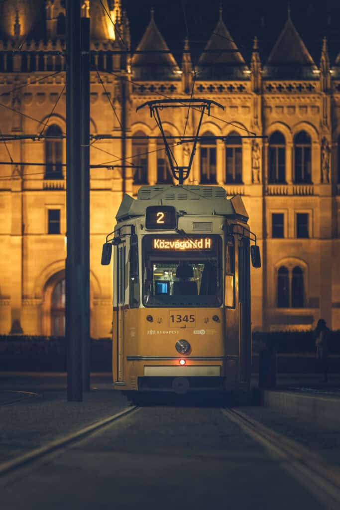 Yellow Budapest tram at night on Tram 2, headlights glowing as it stops on tracks with the illuminated Hungarian Parliament building blurred in the background.