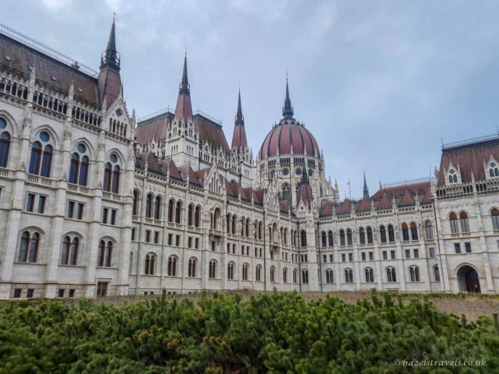 Hungarian Parliament Building in Budapest during daylight, with white stone facade, red-tiled roofs, Gothic spires and central dome rising above green shrubs under an overcast sky.