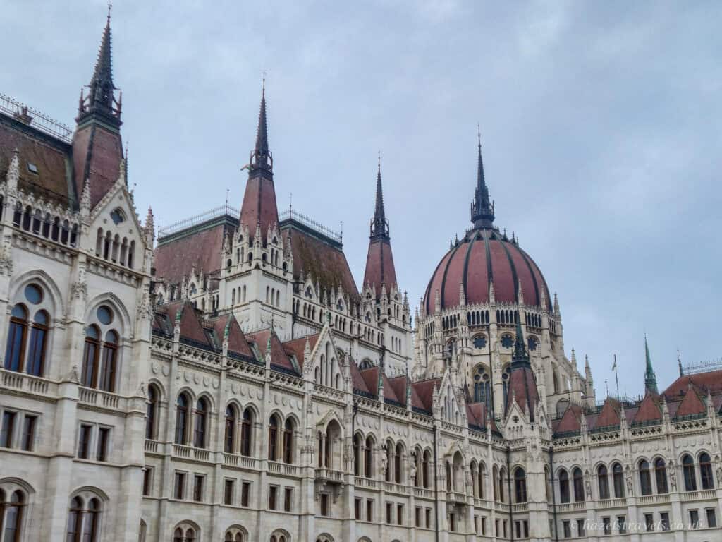 Hungarian Parliament Building in Budapest during daylight, with ornate white stone facade, red-tiled roofs, Gothic spires and a large central dome under a pale grey sky.