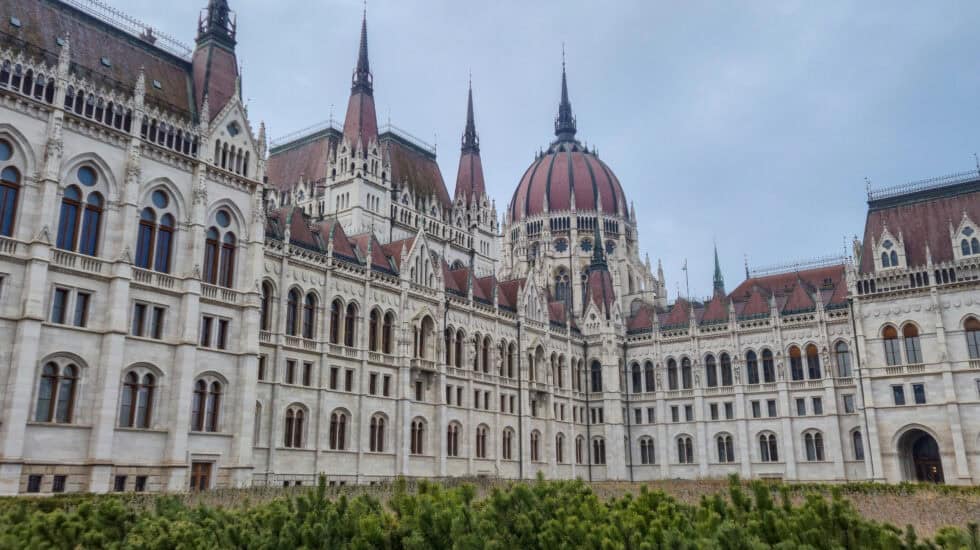 Hungarian Parliament Building in Budapest during daylight, with white stone facade, red-tiled roofs, Gothic spires and central dome rising above green shrubs under an overcast sky.