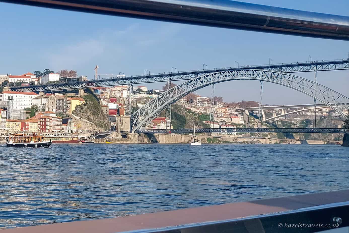 View of the Dom Luís I Bridge spanning the River Douro in Porto, seen from a boat on the water, with colourful hillside buildings, boats, and a clear blue sky in the background.
