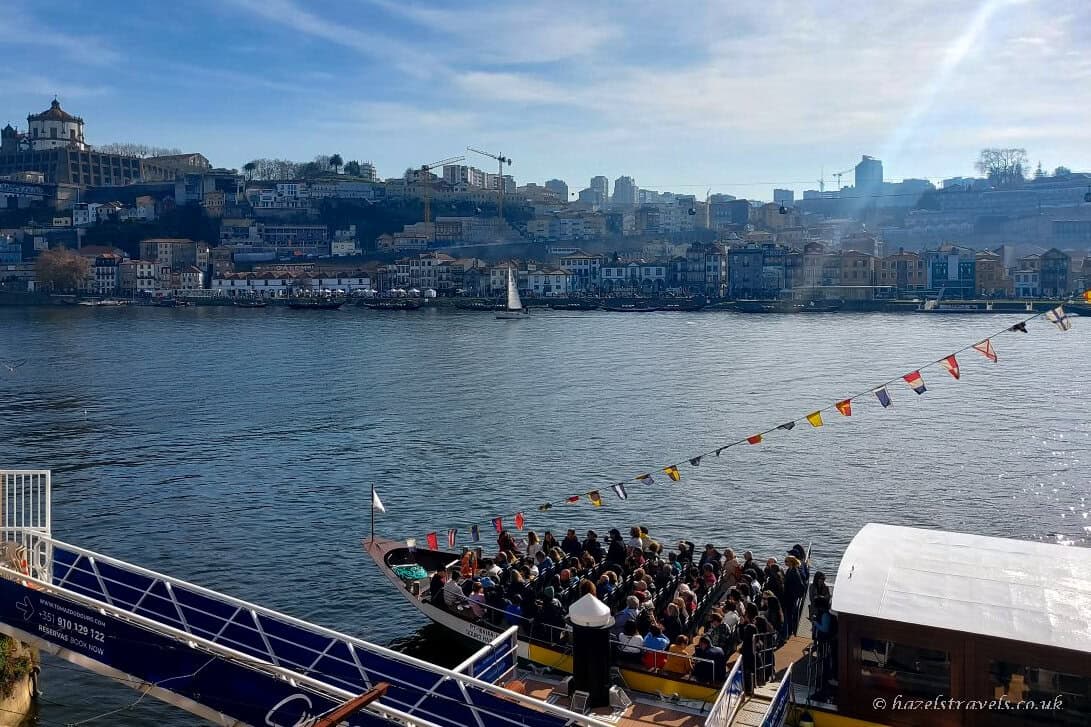 Boat filled with passengers departing on the River Douro in Porto, with colourful flags strung above the deck and the hillside cityscape and riverside buildings visible across the water in bright daylight.