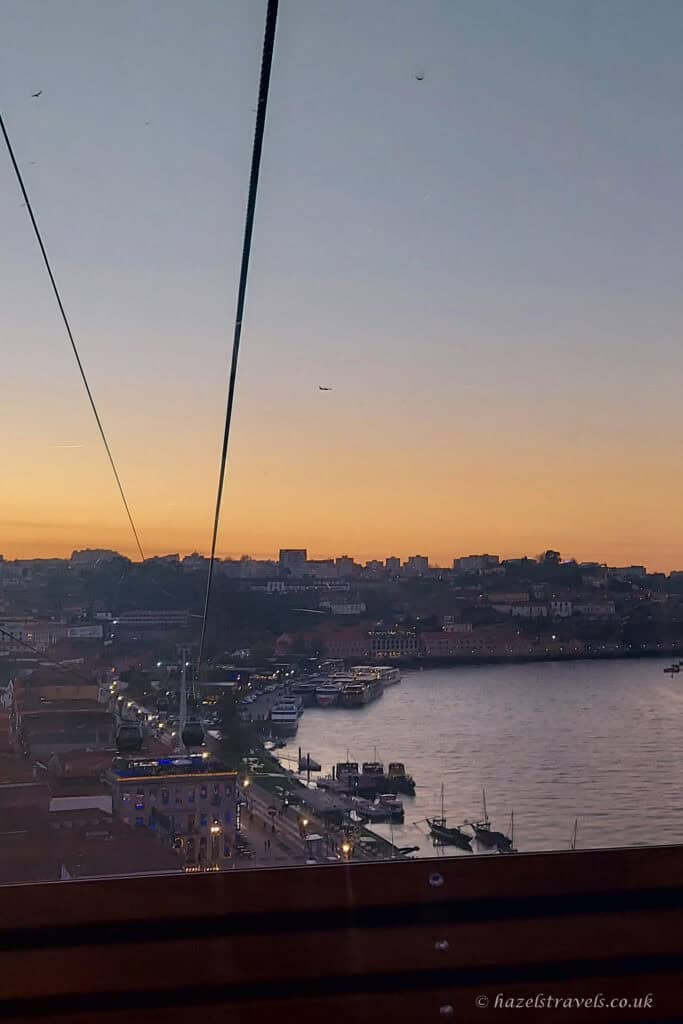 Sunset view over the River Douro in Porto, seen from a cable car, with boats along the riverbank, city buildings on the hillside, and the sky fading from orange to blu
