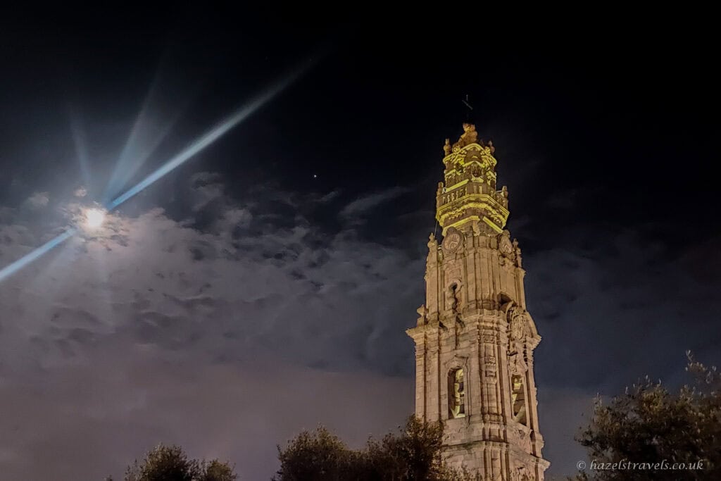 Night-time view of the Clérigos Tower in Porto illuminated against a dark sky, with the moon glowing through clouds and light beams stretching across the scene.