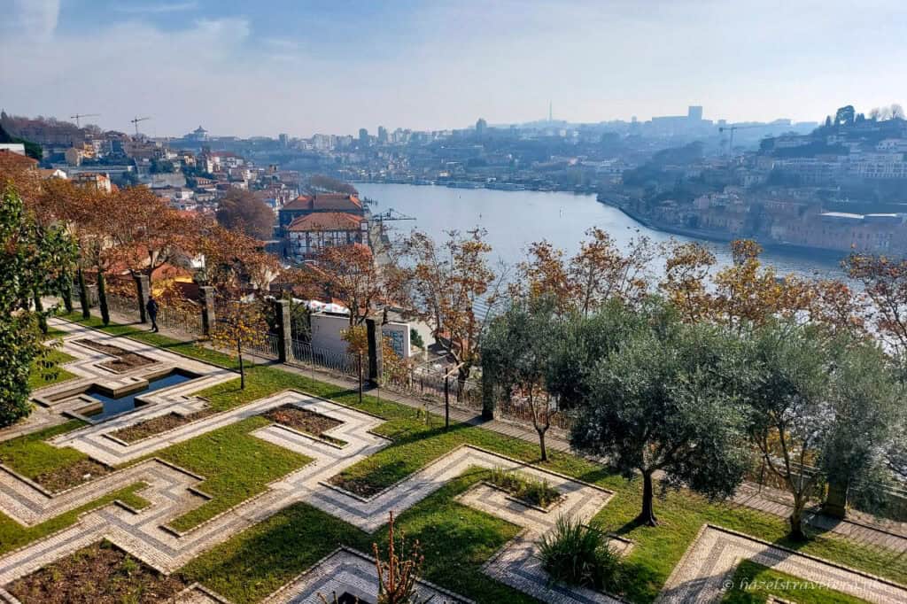 View over the Douro River and Porto skyline from Crystal Palace Gardens, with geometric garden paths, trees, and autumn foliage in the foreground under a hazy blue sky.