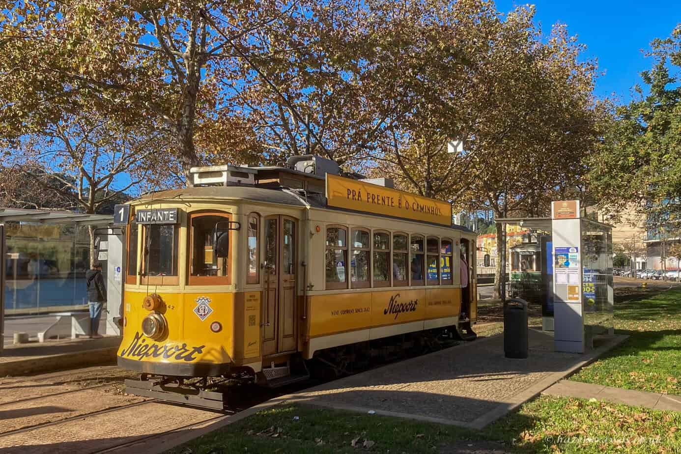 Vintage yellow Porto tram on Line 1 (Infante) stopped at a tree-lined tram stop by the River Douro, with autumn leaves overhead and clear blue skies.