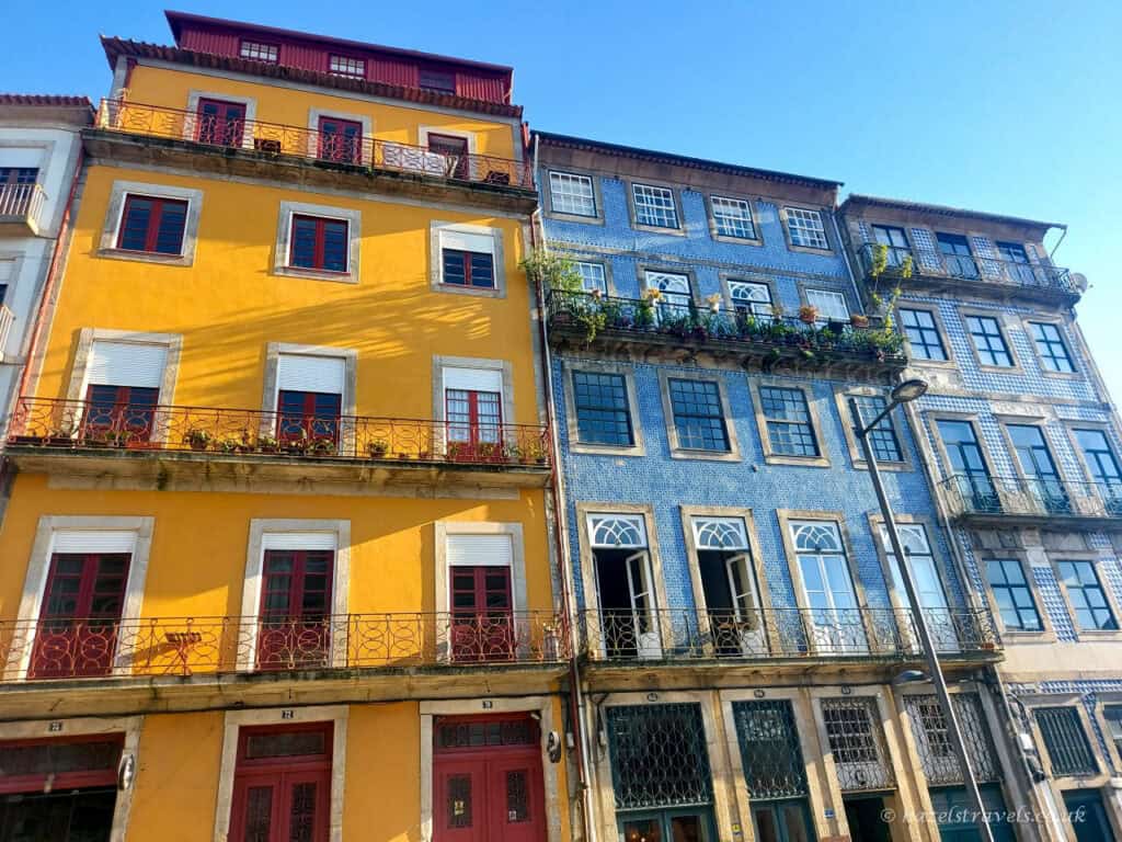 Colourful historic buildings in Porto, with one painted yellow and the other covered in blue-and-white azulejo tiles, featuring wrought-iron balconies and tall windows under a clear blue sky.