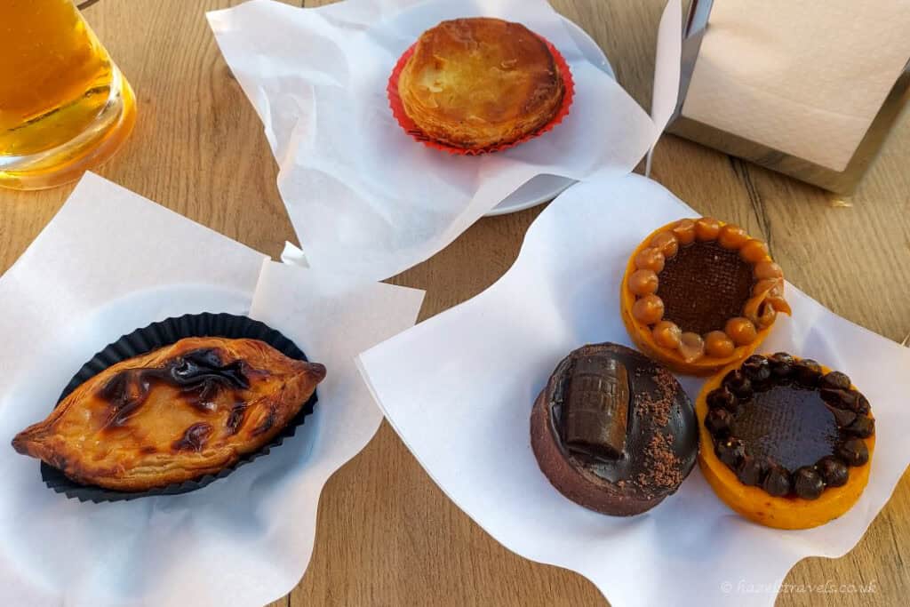 An assortment of Portuguese pastries on a wooden table, including custard tarts, a chocolate tart, and a caramel-topped tart, each served on white paper, with a glass of beer partially visible nearby.
