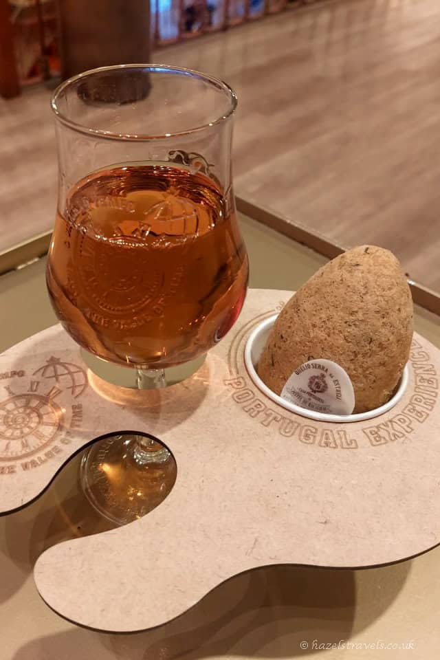 Glass of amber-coloured port wine served on a tasting tray in Porto, accompanied by a small cork-shaped snack and branded mat referencing Portugal’s wine heritage.