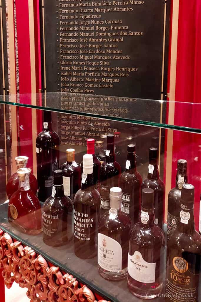 A glass display case filled with bottles of aged port wine, with labels such as 10-year-old tawny visible, and a dark wall behind listing names in gold lettering.