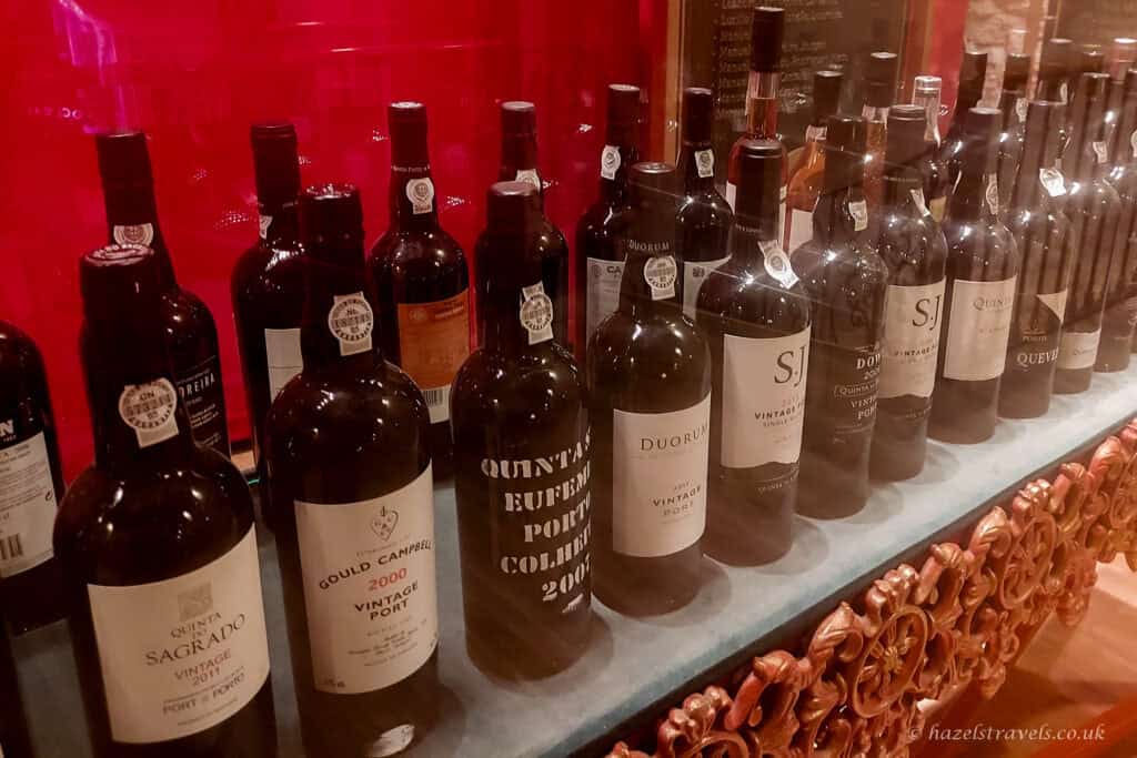 Row of vintage port wine bottles displayed behind glass in a Porto wine cellar, with labels showing different quintas and vintages against a deep red background.