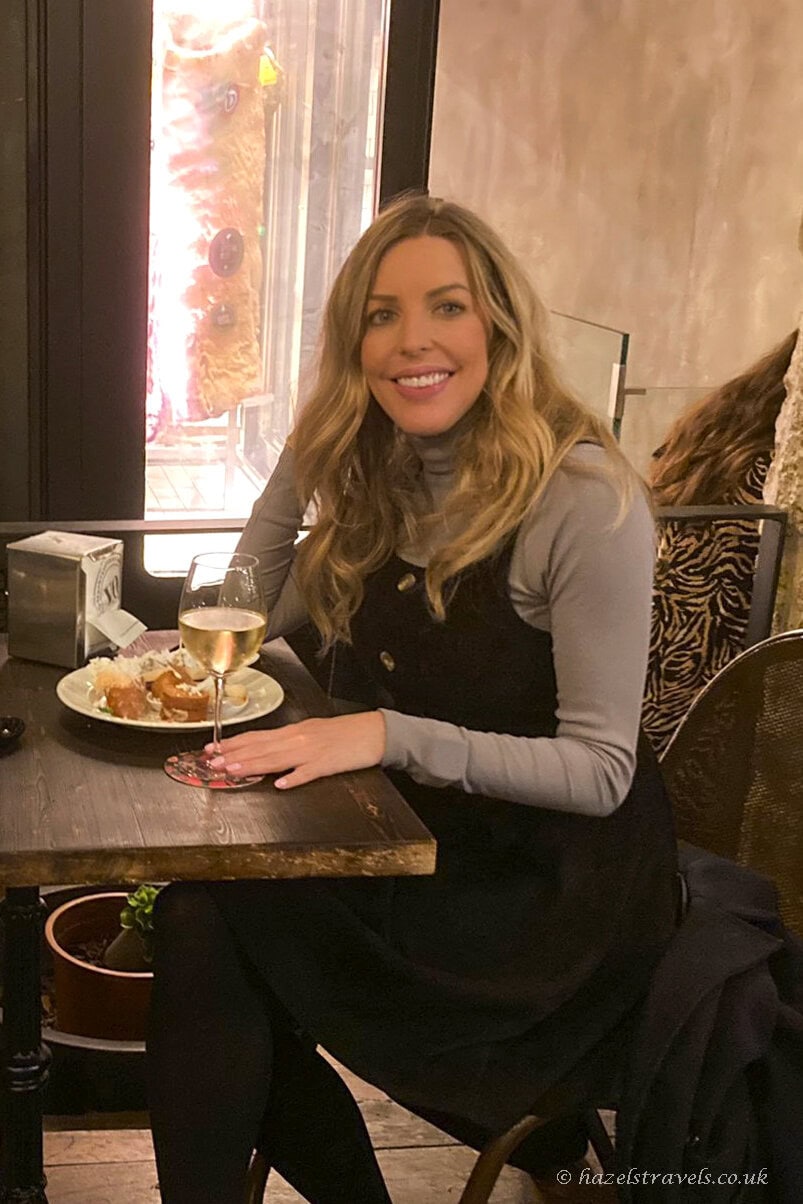 A woman sits at a small restaurant table in Porto, smiling at the camera while holding a glass of white wine, with a plate of tapas and warm indoor lighting around her.