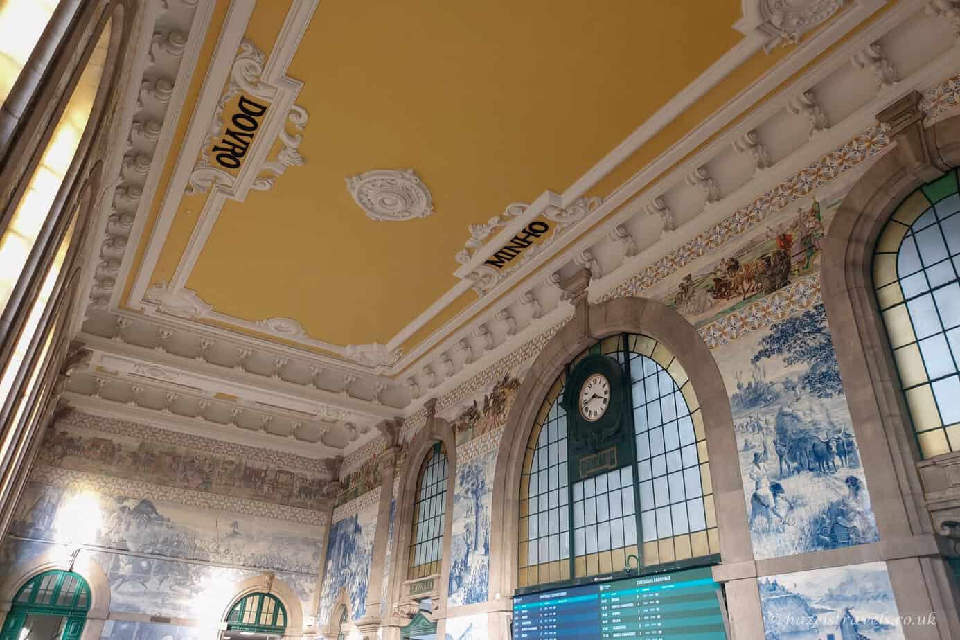 Interior of São Bento railway station in Porto, showing ornate blue-and-white azulejo tile panels, decorative plasterwork, and a large arched window beneath a yellow ceiling.
