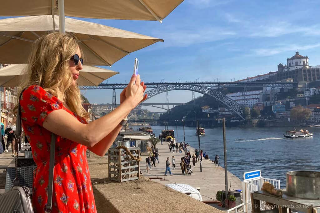 A woman in a red dress stands at a riverside café in Porto, taking a photo on her phone under sun umbrellas, with the River Douro, Dom Luís I Bridge, boats, and hillside buildings in the background.