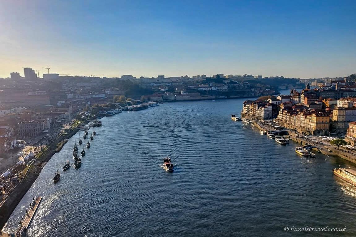 Wide view over the River Douro in Porto, with boats moving along the water and the colourful Ribeira district lining the right-hand riverbank under clear blue skies.