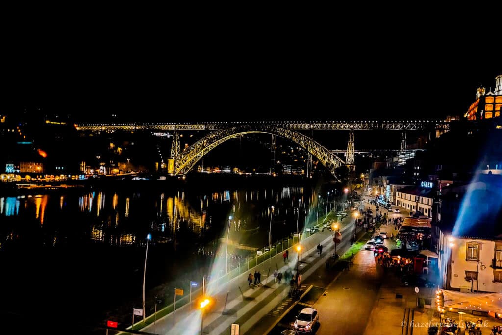 Night-time view over the River Douro in Porto from a rooftop bar, with the Dom Luís I Bridge lit up in gold, reflections shimmering on the water, and the riverside street and cafés glowing below.