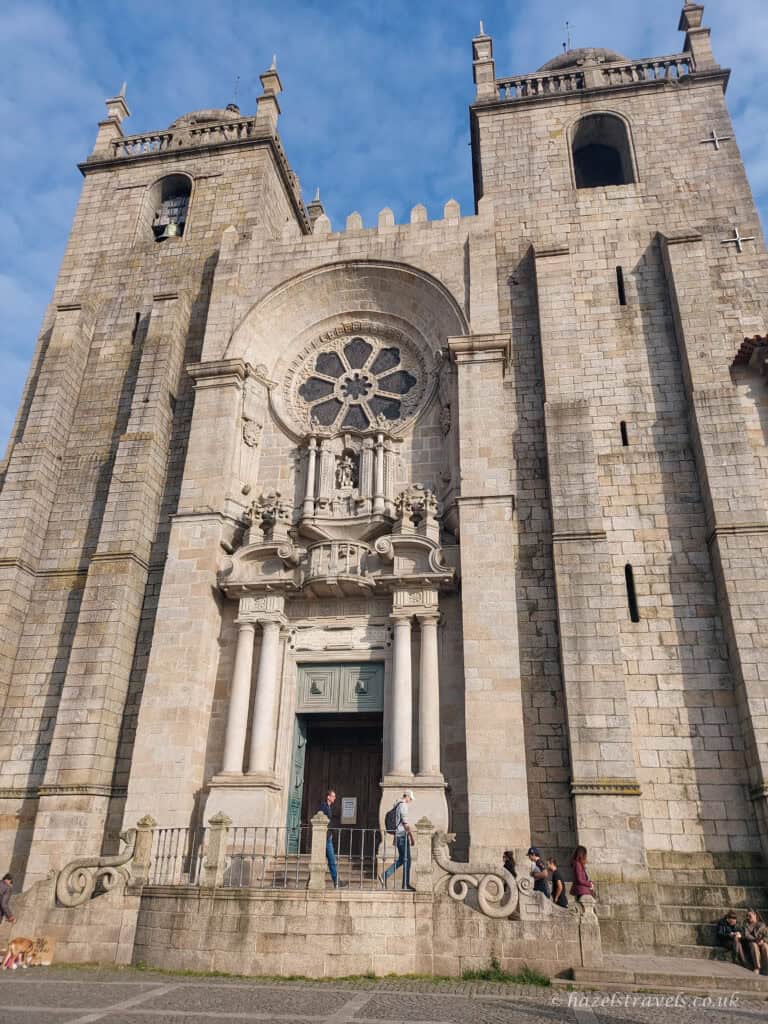 Front view of Porto Cathedral with its twin stone towers, ornate baroque doorway, and large rose window, with people standing on the steps below under a blue sky.