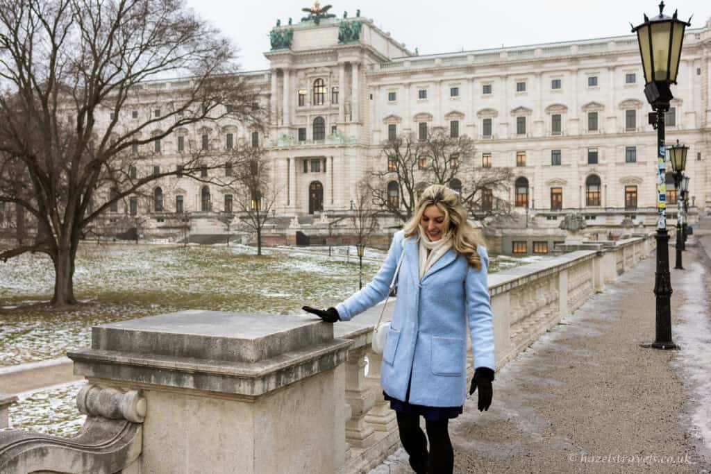 Woman walking along a stone balustrade in winter with the Hofburg Palace in Vienna in the background, bare trees and light snow on the ground.