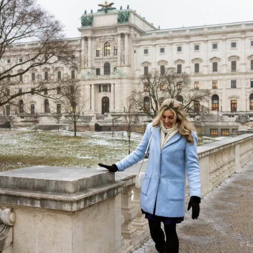 Woman walking along a stone balustrade in winter with the Hofburg Palace in Vienna in the background, bare trees and light snow on the ground.