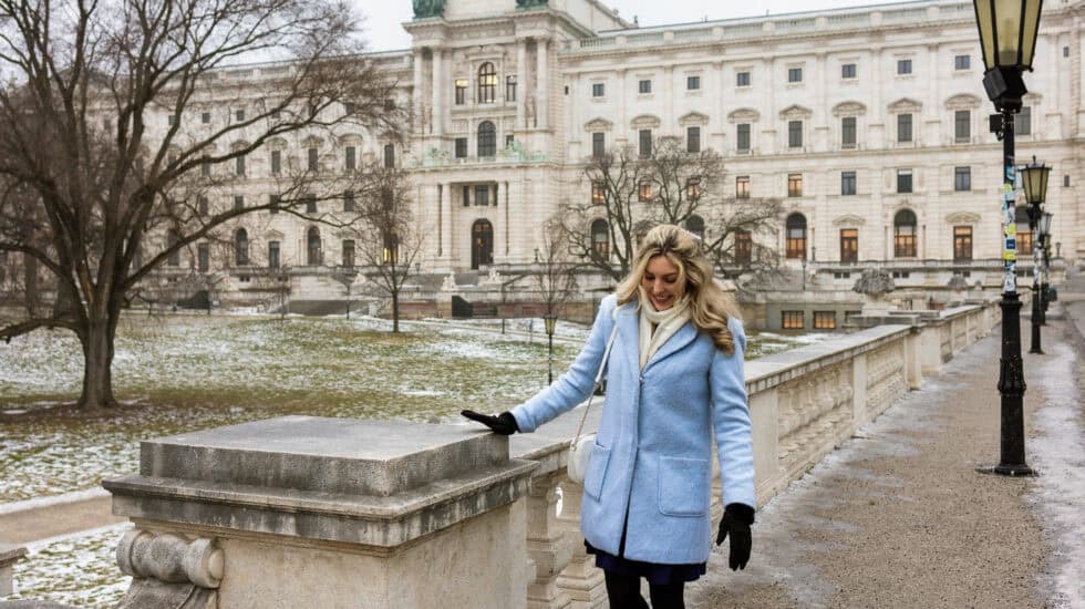 Woman walking along a stone balustrade in winter with the Hofburg Palace in Vienna in the background, bare trees and light snow on the ground.