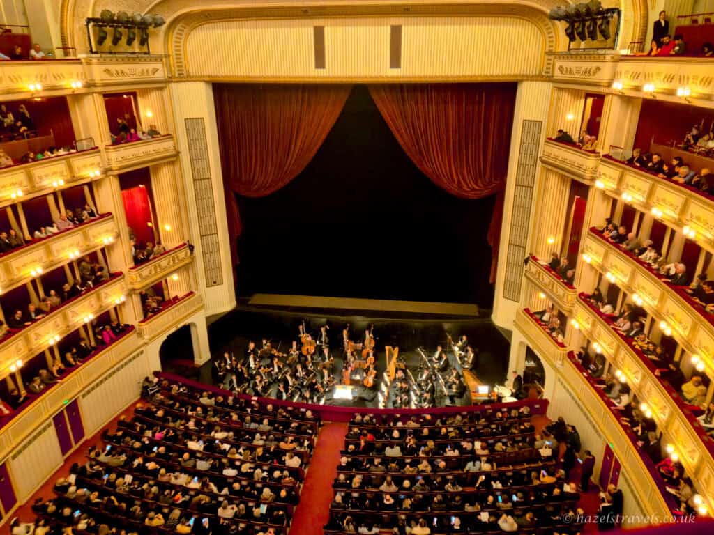 Interior of Vienna State Opera with orchestra performing on stage beneath red velvet curtains, surrounded by ornate cream and gold balconies filled with seated audience members.
