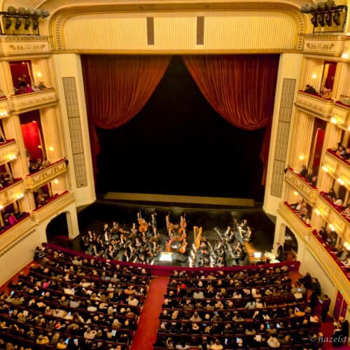 Interior of Vienna State Opera with orchestra performing on stage beneath red velvet curtains, surrounded by ornate cream and gold balconies filled with seated audience members.