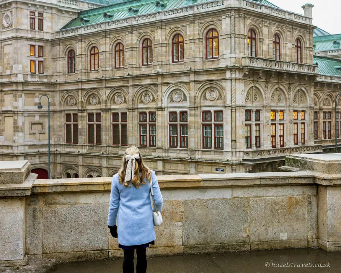 Woman with long blonde hair wearing a light blue coat, black tights and ankle boots, standing on a stone terrace facing the ornate façade of the Vienna State Opera, with its pale stone arches and green copper roof visible above.