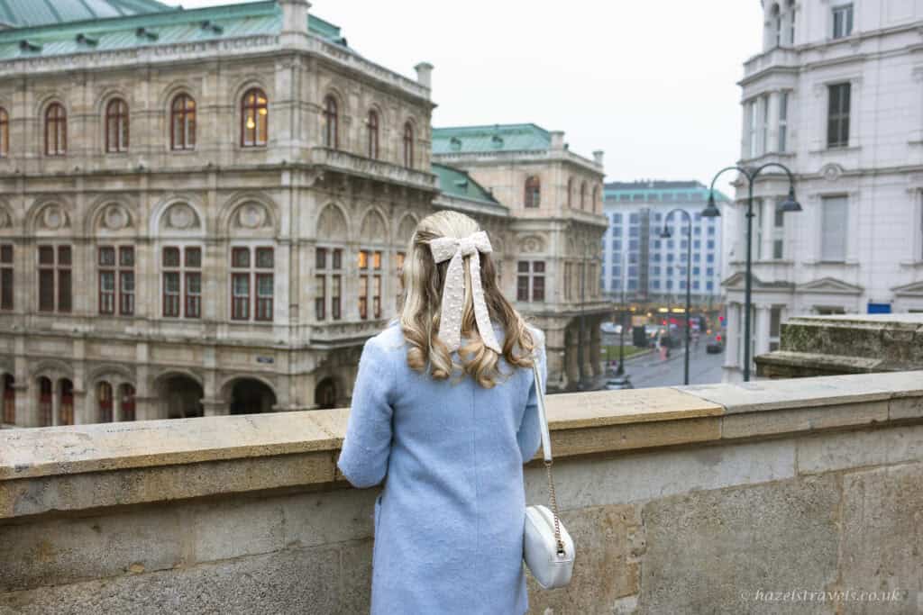 A woman in a pale blue coat stands with her back to the camera, looking out over the Vienna State Opera from a stone terrace on an overcast day, with historic buildings and a city street below.