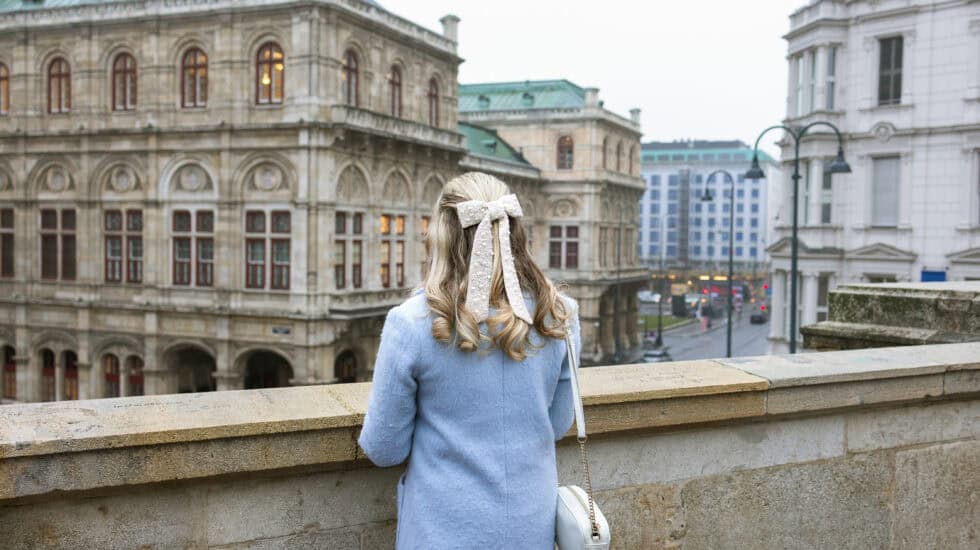 A woman in a pale blue coat stands with her back to the camera, looking out over the Vienna State Opera from a stone terrace on an overcast day, with historic buildings and a city street below.