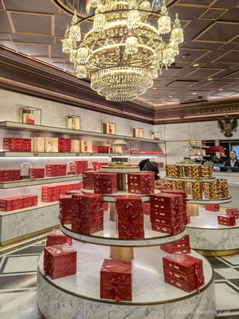 Elegant interior of a Vienna coffee house with crystal chandeliers, marble display tables stacked with red cake boxes, and traditional patisserie counters in the background.
