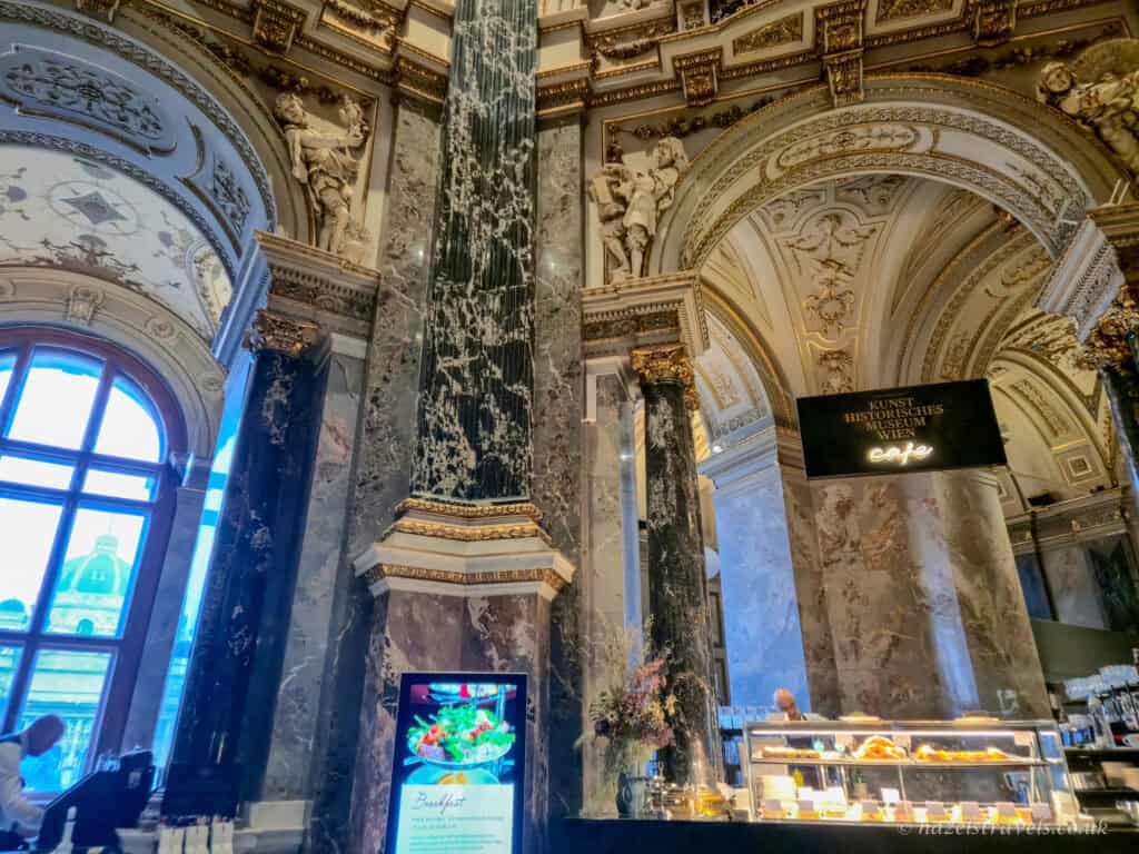 Grand interior of a Vienna coffee house inside the Kunsthistorisches Museum, with marble columns, ornate arches, chandeliers, and a café counter beneath towering ceilings.