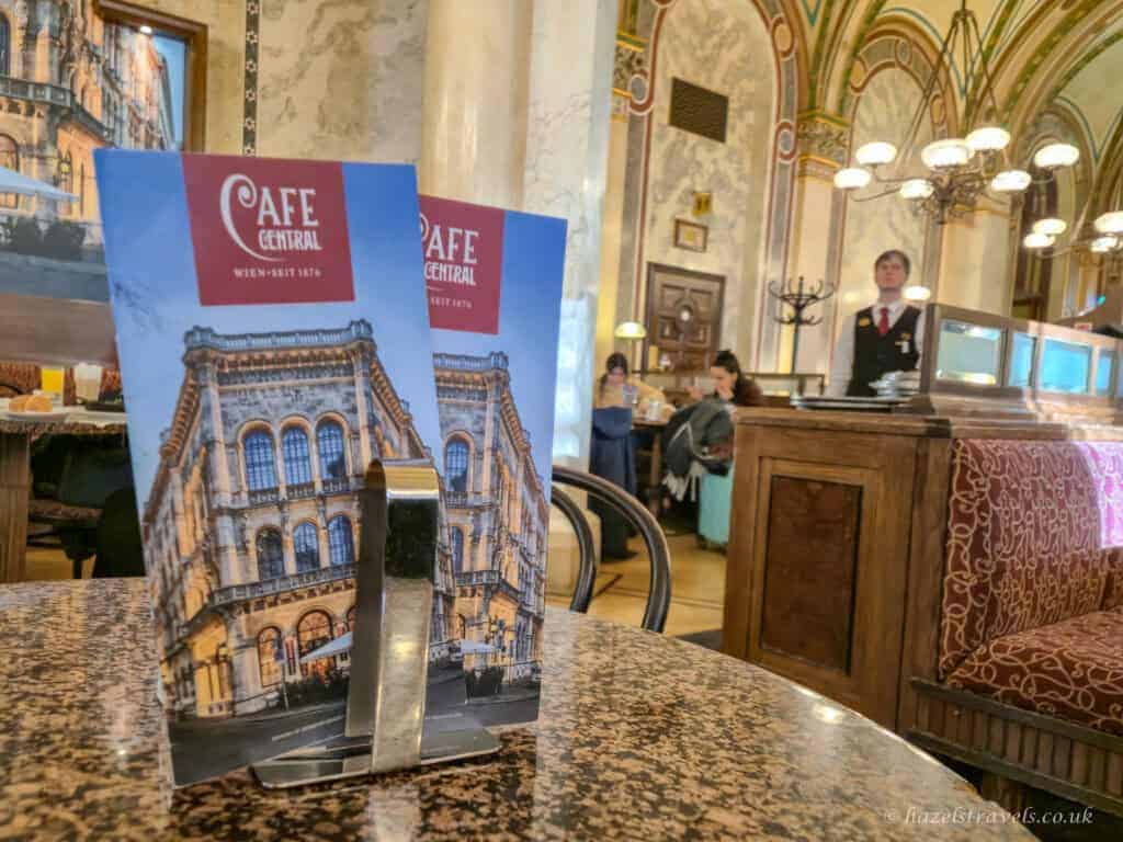 Menu stand on a marble table inside Café Central, one of the most famous Vienna coffee houses, with ornate ceilings and traditional interior in the background.