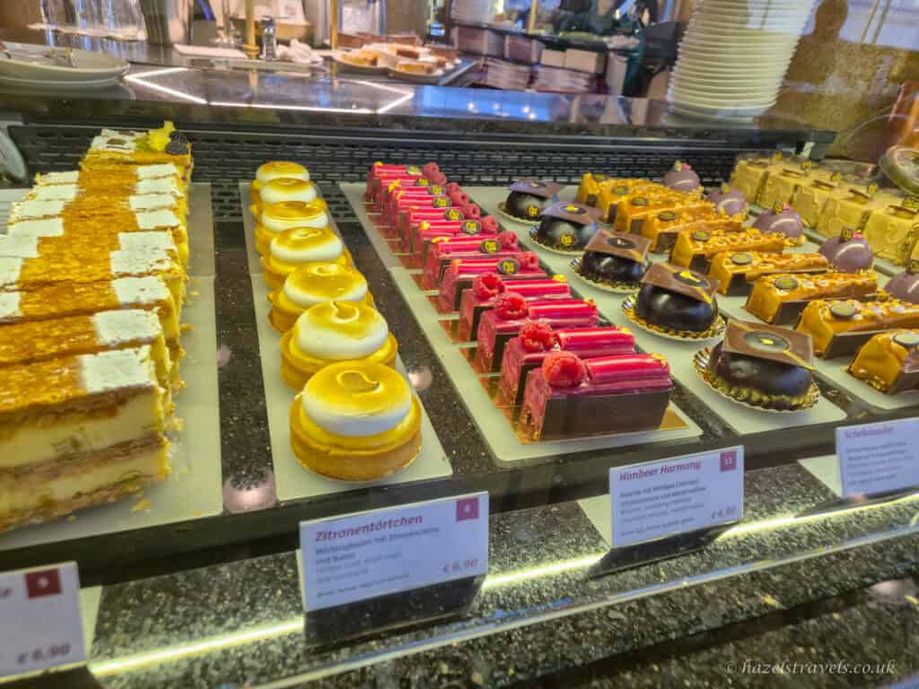 Glass display case filled with neatly arranged pastries and cakes at Café Central, including lemon tarts, chocolate domes, and raspberry-topped slices with small price labels in front.