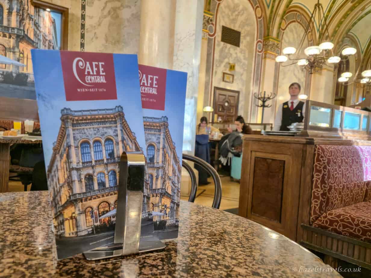 Menu stand on a marble table inside Café Central, one of the most famous Vienna coffee houses, with ornate ceilings and traditional interior in the background.