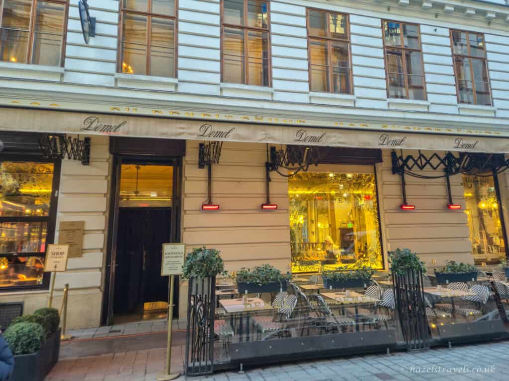 Exterior of Demel café in Vienna, with cream-coloured façade, gold-lit display windows, outdoor seating, and the Demel name on the awning above the entrance.