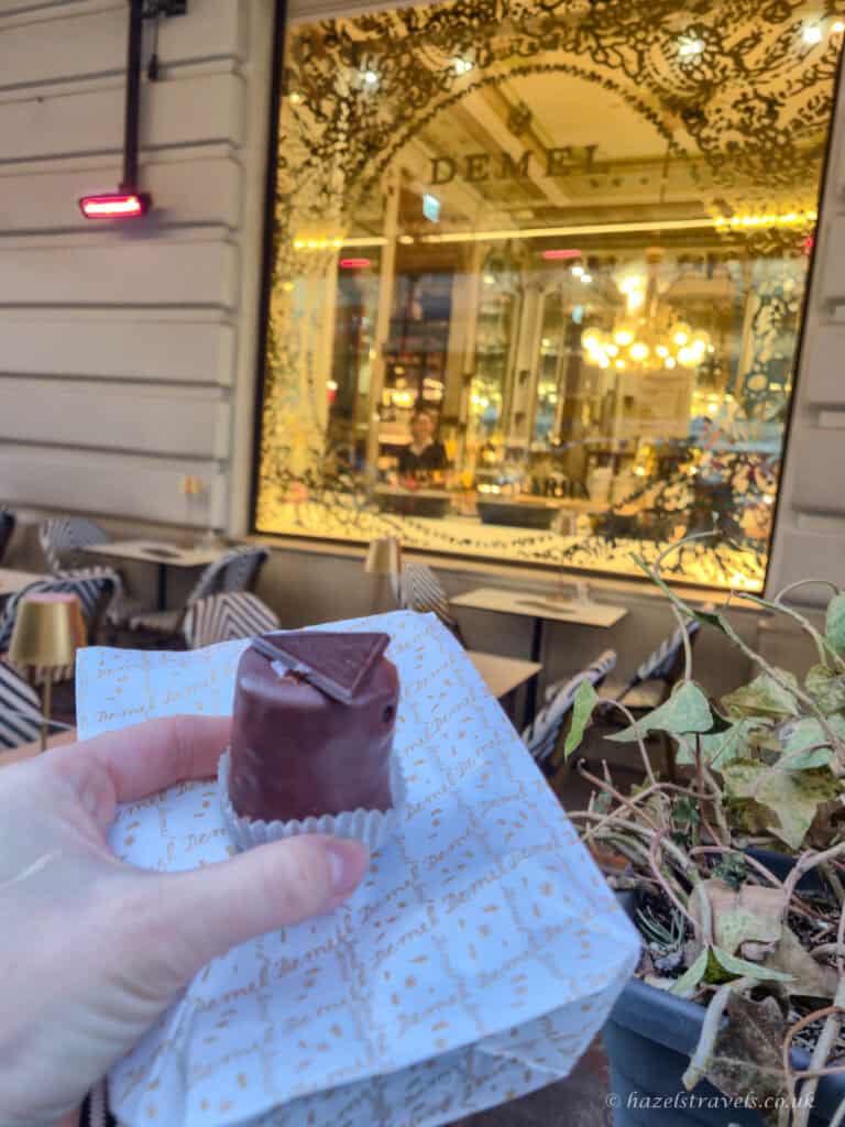 Hand holding a small chocolate-covered cake outside Demel in Vienna, with the café’s gold-lit window and outdoor tables in the background.