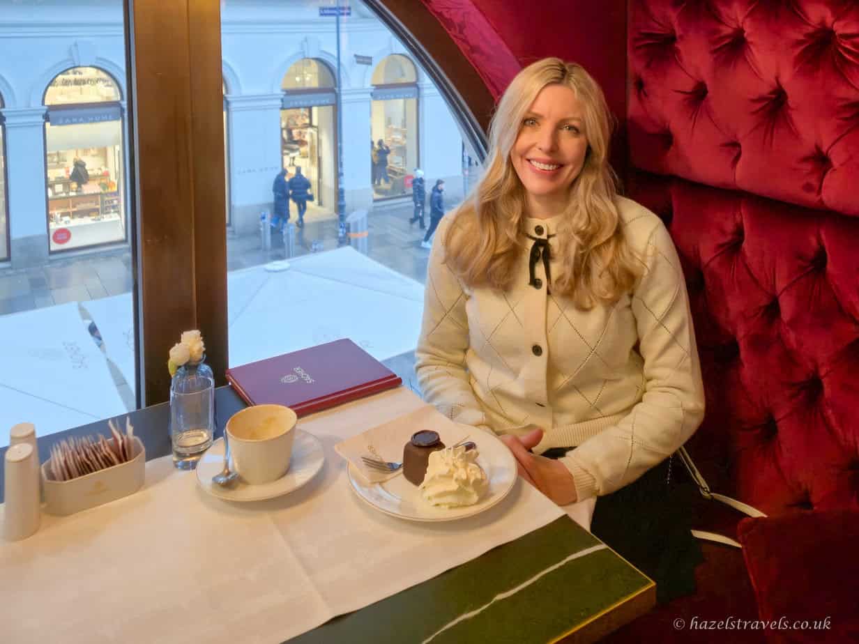 Hazel sitting at a marble table inside a traditional Vienna coffee house, with a slice of chocolate cake and whipped cream, a melange, and a red chandelier overhead by the window.