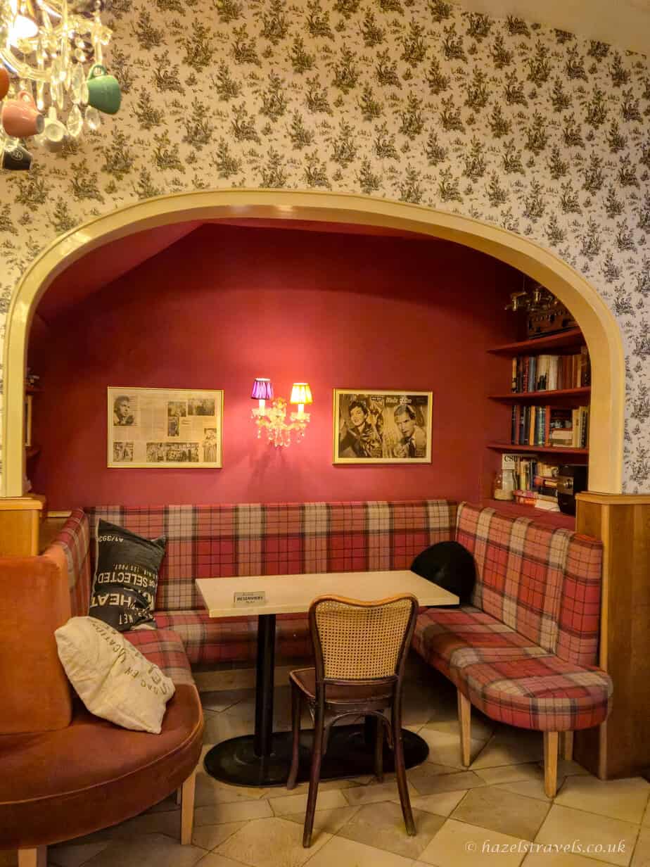 Cosy seating area inside a Vienna café, with red walls, patterned wallpaper, plaid bench seating, framed artwork, and a small table beneath a chandelier with hanging cups.