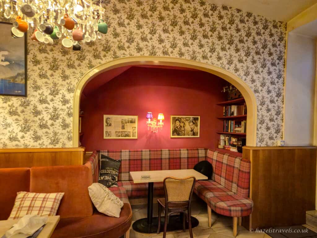 Cosy seating area inside a Vienna café, with red walls, patterned wallpaper, plaid bench seating, framed artwork, and a small table beneath a chandelier with hanging cups.